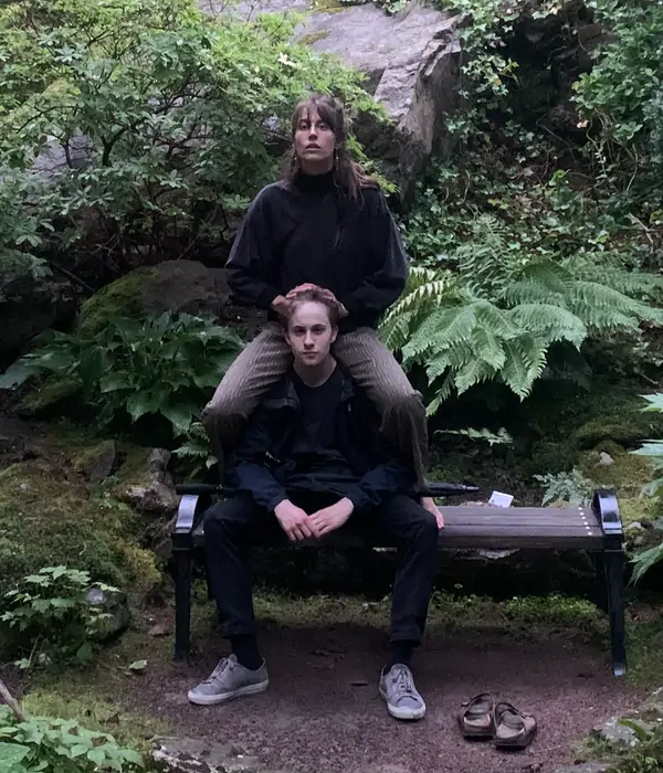 Två personer, Vivekas barn sitter på en bänk omgiven av grönska och fjädrande växter.