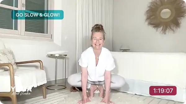 Viveka sitter på en ljus matta i ett välbelyst rum och ler mot kameran. Hon är i en yogaposition med benen brett isär och händerna placerade på golvet framför sig. Hon bär en vit, löst sittande outfit. I det övre vänstra hörnet finns texten *