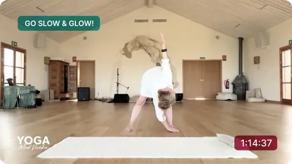 Viveka sitter på en ljus matta i ett välbelyst rum och ler mot kameran. Hon är i en yogaposition med benen brett isär och händerna placerade på golvet framför sig. Hon bär en vit, löst sittande outfit. I det övre vänstra hörnet finns texten *