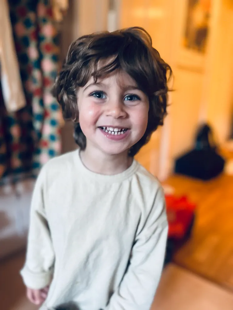 En liten pojke med lockigt, brunt hår och blå ögon ler brett. Han bär en ljus, långärmad tröja. Bakgrunden visar en hemmet med suddiga färger och olika föremål.
